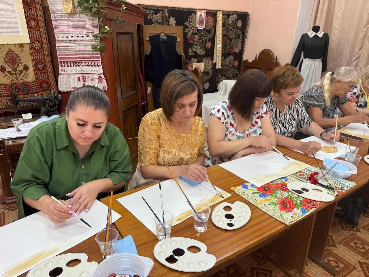 Працівниця музею «Степова Україна» у золотистій сукні та переселенки з Херсонщини зосереджено малюють за довгим дерев'яним столом під час майстер-класу з кавового живопису. На передньому плані розкладені палітри з кавовим розчином, аркуші паперу та пензлі, а на тлі кімнати представлено автентичні вишиті рушники й ткані килими.