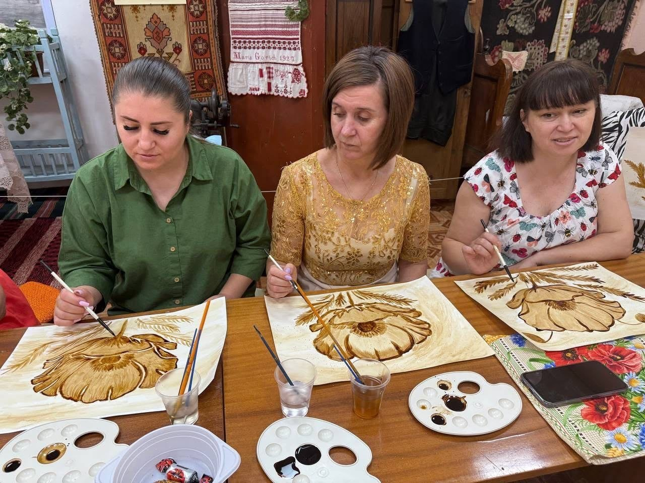 Працівниця музею у золотистому вбранні та дві переселенки з Херсонщини малюють великі квітки маку в техніці кавового живопису, сидячи за дерев'яним столом. Перед ними розкладені палітри з кавовим розчином різних відтінків, пензлі та склянки з водою, а на тлі музею «Степова Україна» видно вишиті рушники.