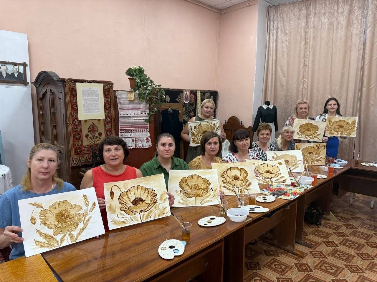 Групове фото переселенок із Херсонщини та працівниці музею «Степова Україна» в золотистому вбранні. Учасниці заходу сидять і стоять уздовж довгого столу, тримаючи перед собою готові малюнки великих маків, виконані у техніці кавового живопису.
