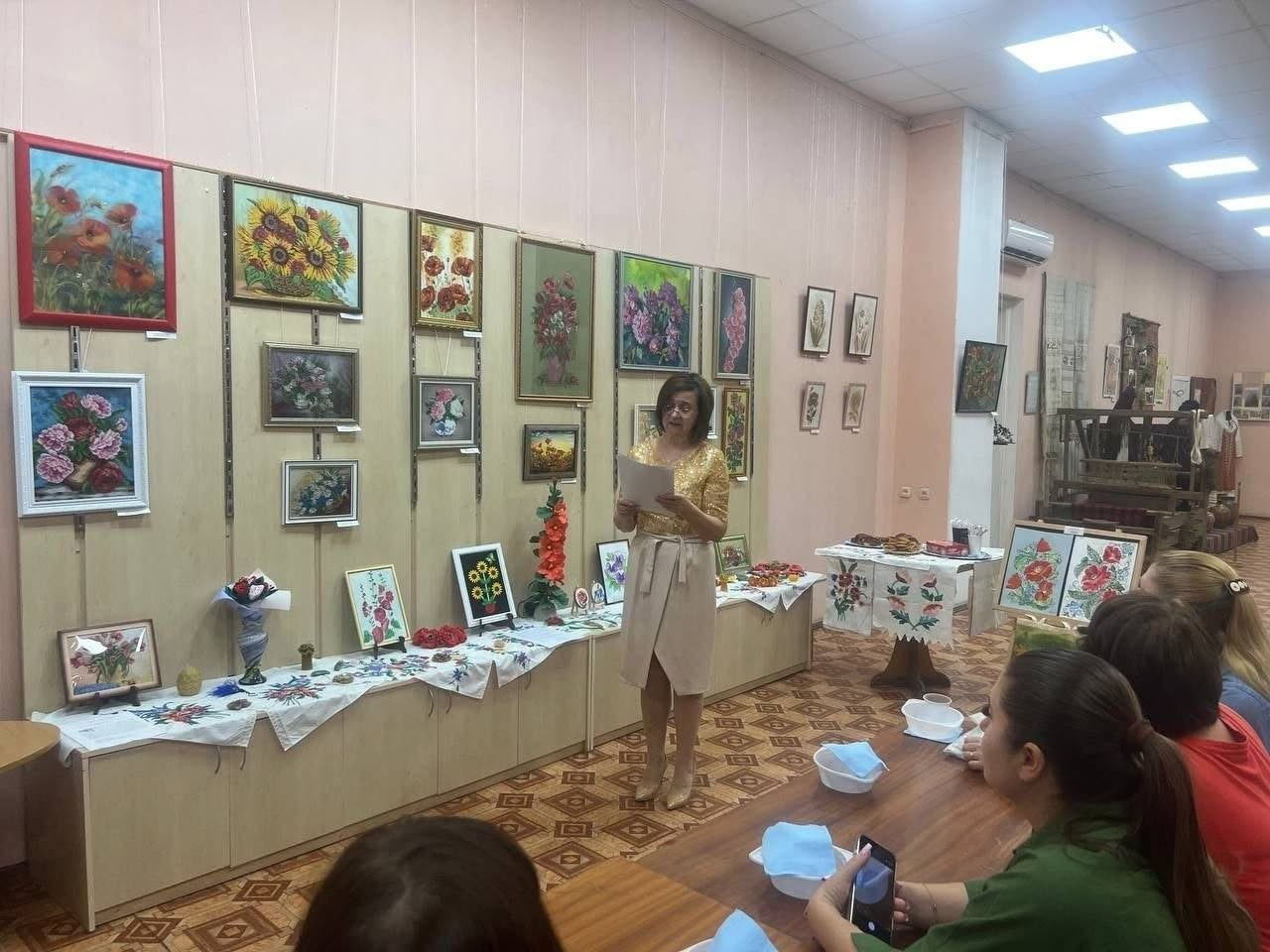 Виступ ведучої заходу перед переселенками з Херсонщини в музеї «Степова Україна». Жінка у світлій сукні стоїть по центру зали з аркушем паперу в руках та читає лекцію, поки слухачки сидять за столом праворуч. На стінах та столах навколо представлено музейну експозицію: картини з квітами, вишиті рушники, декоративні вироби та макові смаколики.