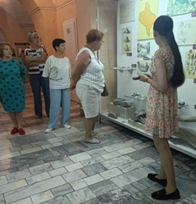Група жінок у музеї слухає екскурсоводку в літній сукні біля вітрини з експонатами та картами на стіні.