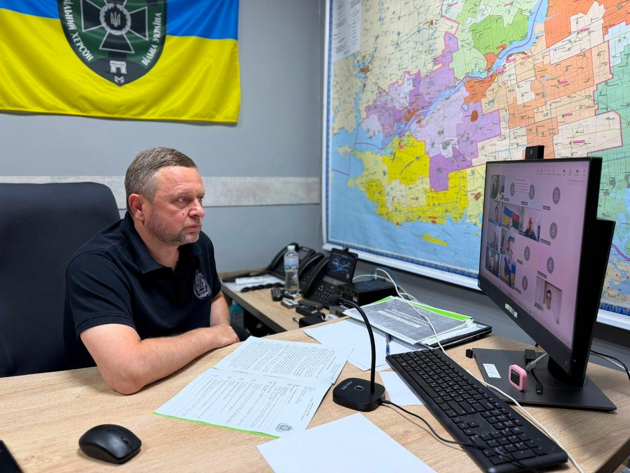 Дмитро Бутрій проводить онлайн-нараду платформи «Діалог влади та бізнесу» за робочим столом у кабінеті, де на стінах розміщено карту Херсонської області та жовто-блакитний прапор з емблемою.