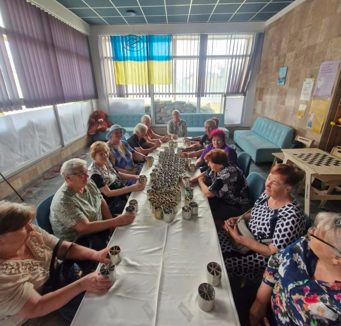 Фотографія ілюструє волонтерську акцію переселенців із Херсонщини у Миколаєві, які виготовляють заготовки для окопних свічок. Літні люди сидять навколо довгого столу, на якому вишикувані десятки підготовлених бляшанок із гофрокартоном, а на вікні кімнати висить прапор України з логотипом БФ «EU4KHERSON».