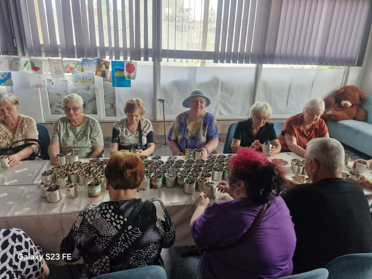 Група літніх переселенців із Херсонщини сидить за столом у Миколаєві та робить заготовки для окопних свічок для Сил оборони. На столі перед волонтерами стоять десятки бляшанок із гофрокартоном, а на вікні позаду них висять дитячі патріотичні малюнки.