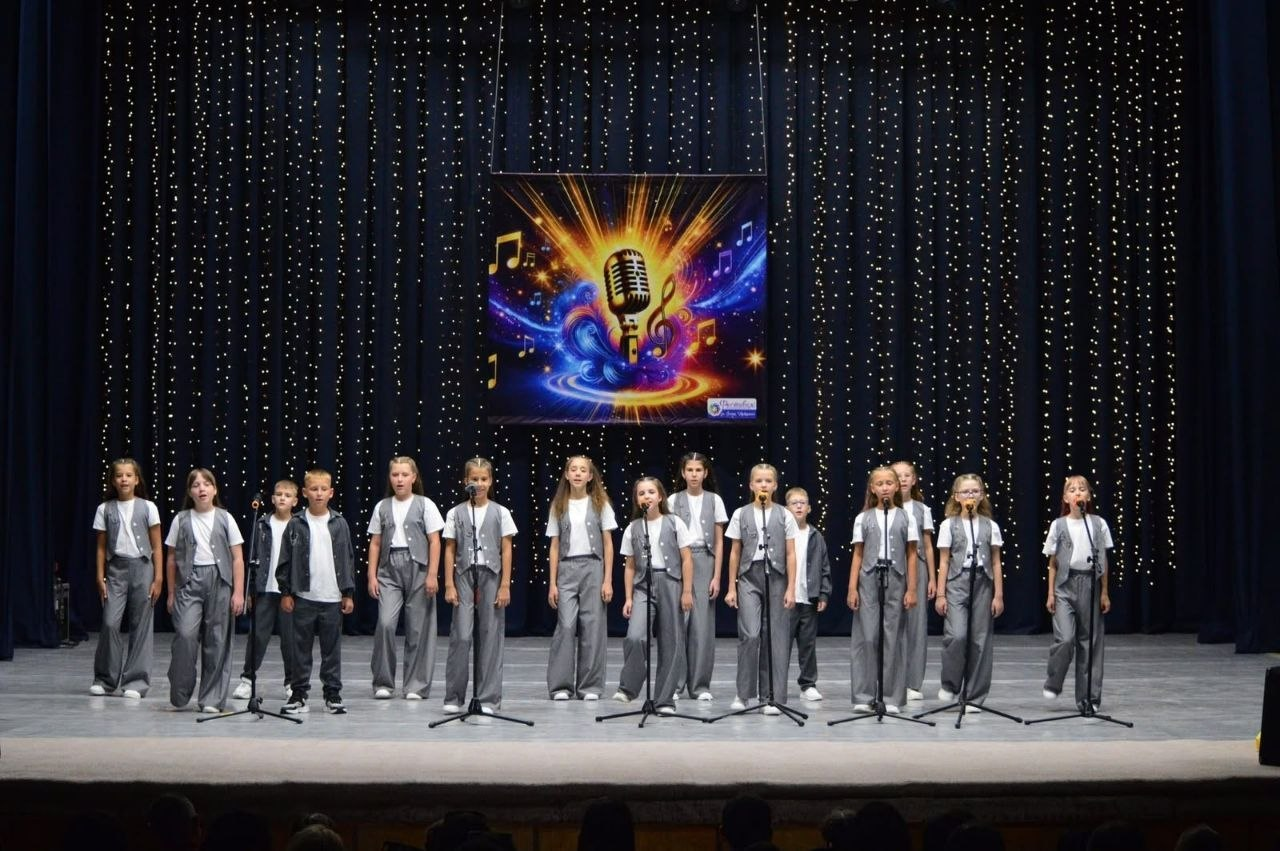 Дитячий вокальний колектив «Mini Star» з вокальної студії «The Best», у складі якого виступають юні переселенці з Верхньорогачицької громади Херсонщини, співає на сцені Міжнародного фестивалю-конкурсу «HIT THE TALENTS 2026» у Львові. Діти у сірих костюмах вишикувані в ряд перед мікрофонами на тлі темної завіси з гірляндами та яскравим екраном із музичною символікою.