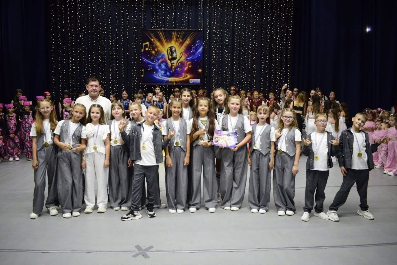 Учасники дитячого вокального колективу «Mini Star» разом із керівником стоять на сцені фестивалю «HIT THE TALENTS 2026» після нагородження. Діти в сірих сценічних костюмах радісно позують із кубком, дипломом переможців та медалями на шиї на тлі інших творчих колективів конкурсу.
