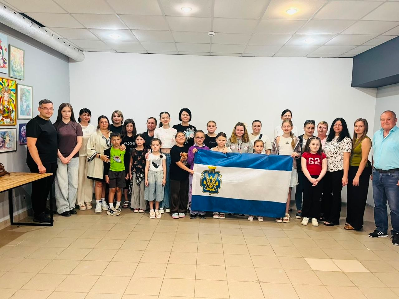 Група дітей-переселенців з Херсонщини та їхніх батьків стоїть у залі Арт-Студії «Дозвілля» у Кривому Розі, тримаючи прапор Херсонської області.
