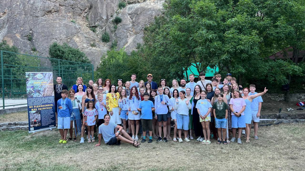 Групове фото учасників програми відпочинку та реабілітації для родин загиблих Захисників і Захисниць України на Закарпатті: дорослі та діти у літньому одязі й вишиванках позують на тлі мальовничої скелі, поруч із банером проєкту «Ми піднімаємося, щоб зцілитися».