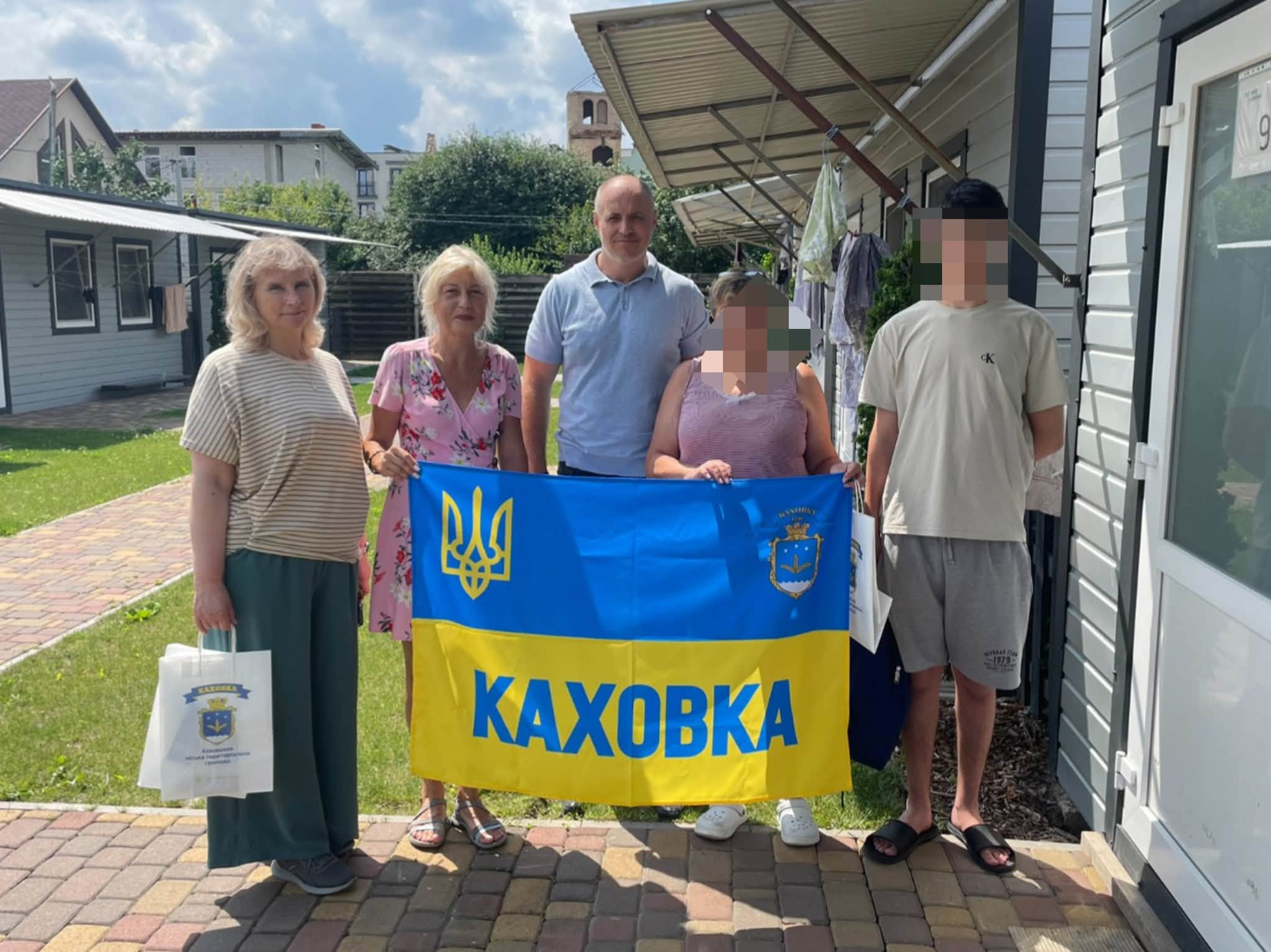 Начальник Каховської МВА Віталій Немерець разом із представниками громади відвідує евакуйовану з тимчасово окупованих територій родину з підлітком, тримаючи синьо-жовтий прапор із написом «Каховка» та передаючи гуманітарні пакунки з речами першої потреби.