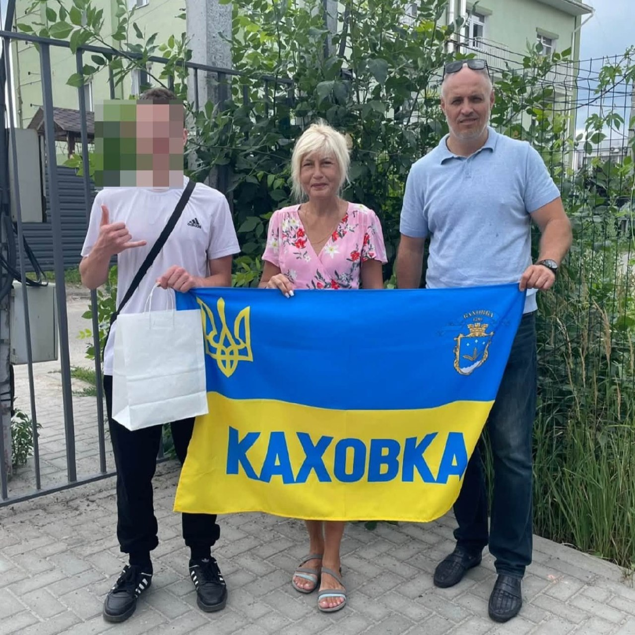 Начальник Каховської МВА Віталій Немерець разом із представницею громади та евакуйованим підлітком позують на вулиці, тримаючи великий синьо-жовтий прапор із гербом та написом «Каховка», а хлопець ліворуч тримає білий пакунок із допомогою.