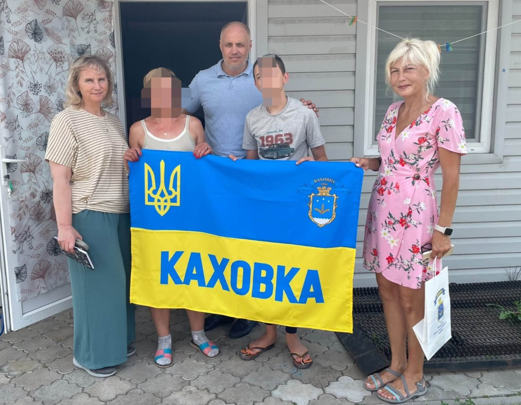 Начальник Каховської МВА Віталій Немерець разом із представницями громади відвідує ще одну евакуйовану родину — жінку та підлітка, тримаючи спільний синьо-жовтий прапор із написом «Каховка» біля входу до будинку.