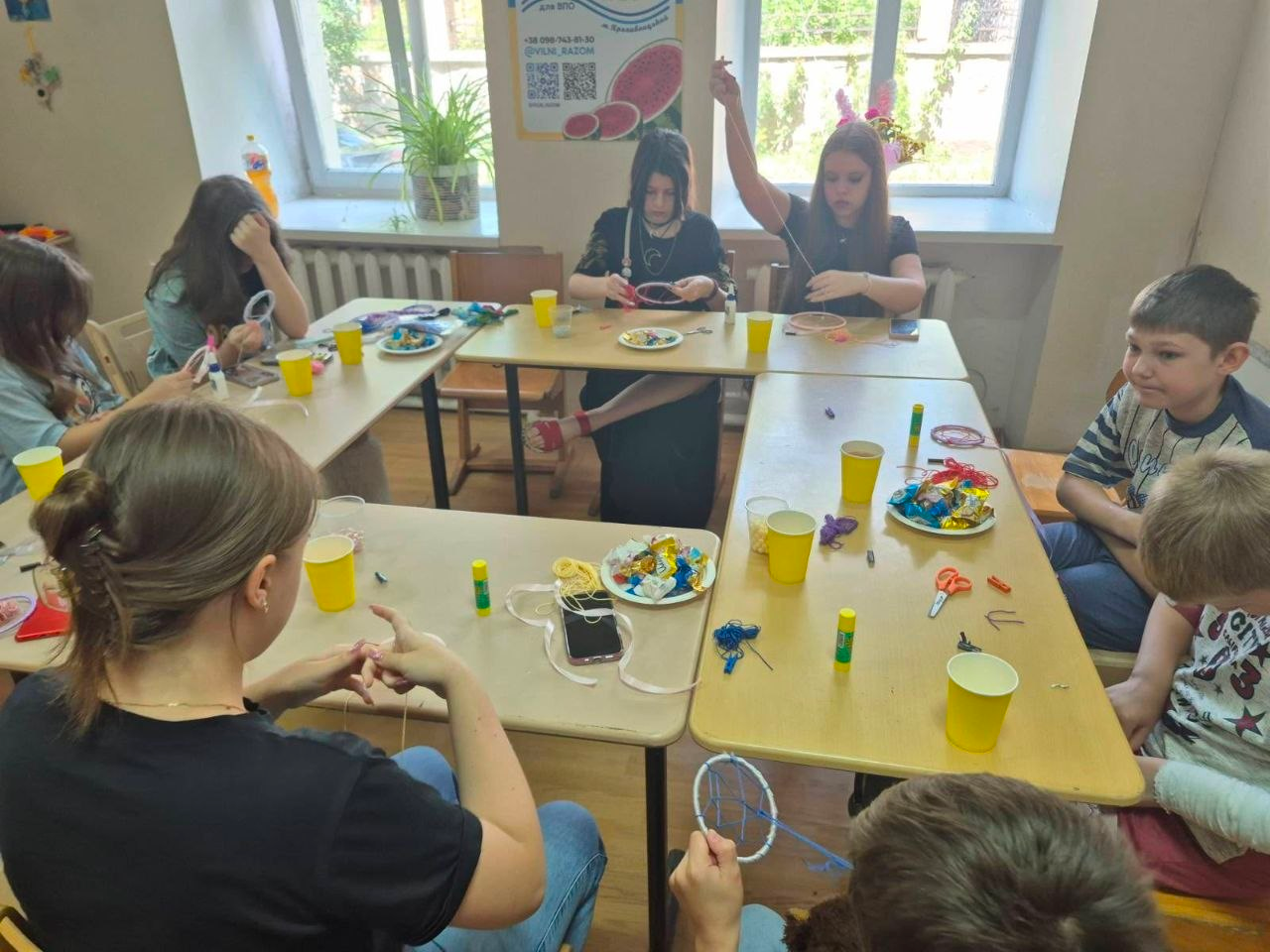 Діти та підлітки-переселенці з Херсонщини плетуть «ловців снів» за великим столом у кропивницькому центрі «Вільні разом»: дівчата на задньому плані натягують нитки на круглі основи, а на столах розкладено матеріали для творчості, ножиці, клей та тарілки із солодощами.