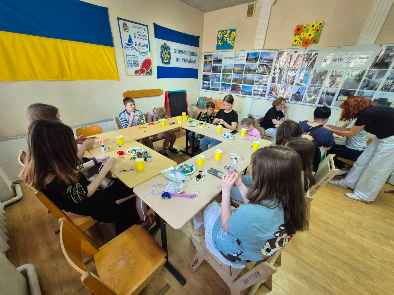 Процес виготовлення оберегів «ловців снів» юними переселенцями з Херсонщини в центрі «Вільні разом» у Кропивницькому: велика група дітей та підлітків працює за П-подібним столом, а на стіні позаду висять державний прапор України, прапор області з написом «Херсонщина це Україна!» та стенди з фотографіями рідного краю.