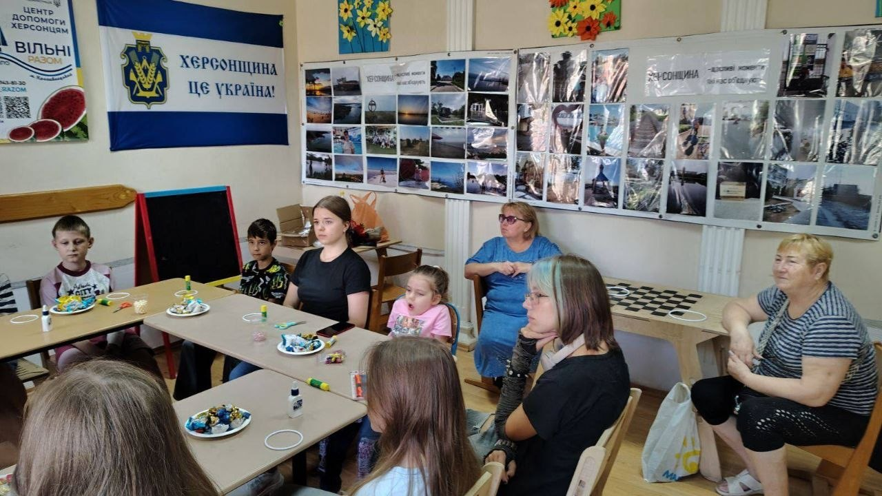 Учасники майстер-класу в кропивницькому центрі «Вільні разом» слухають організаторів перед початком творчої роботи: діти, підлітки та дорослі сидять за столами й на стільцях на тлі прапора області з написом «Херсонщина це Україна!» та фотостендів із краєвидами рідного краю.