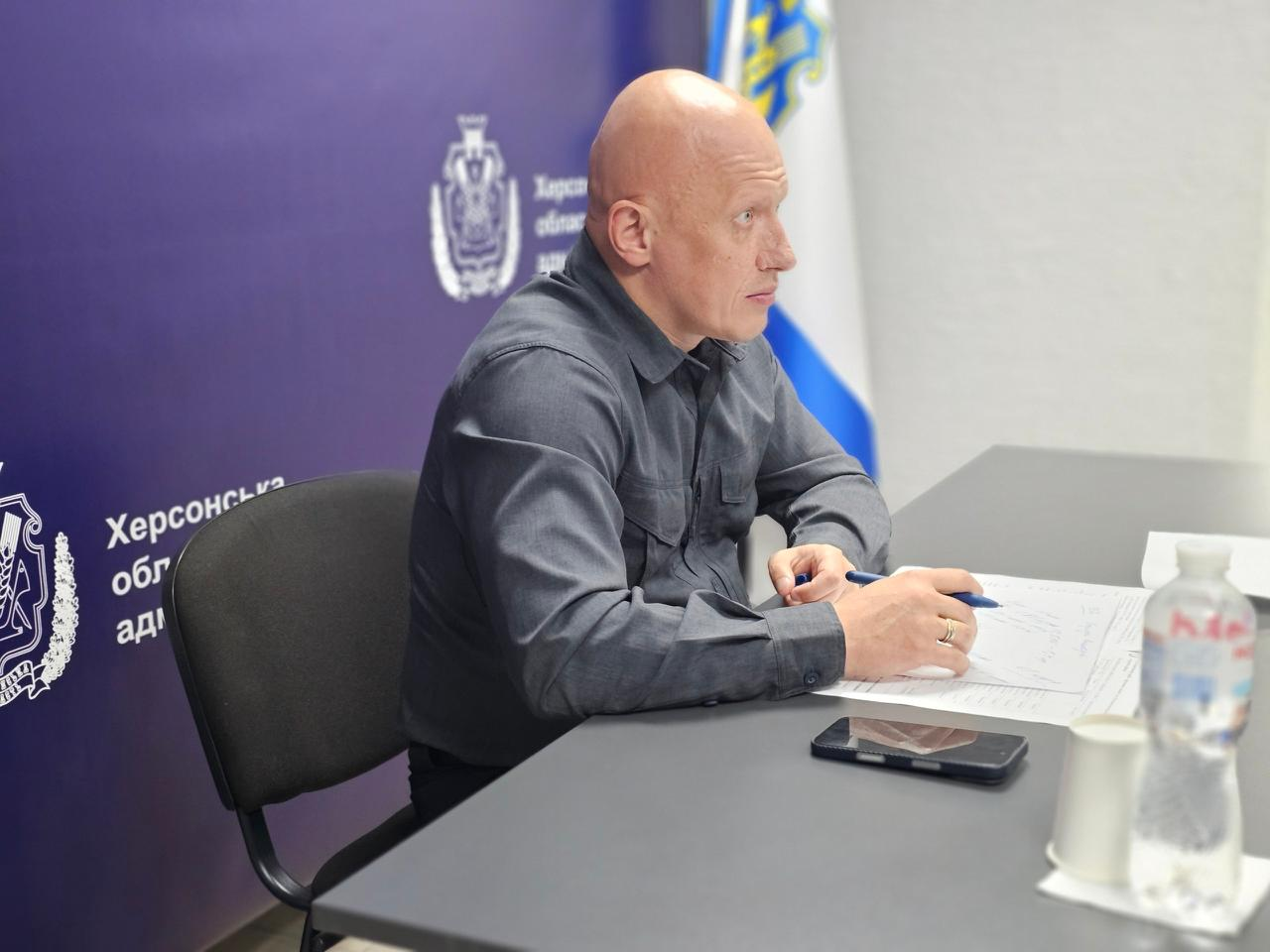 Заступник начальника Херсонської ОВА Євгеній Ігнатенко бере участь в онлайн-нараді Мінрозвитку громад щодо Комплексного плану стійкості, сидячи у профілі за робочим столом із документами та ручкою на тлі офіційного синього банера установи.