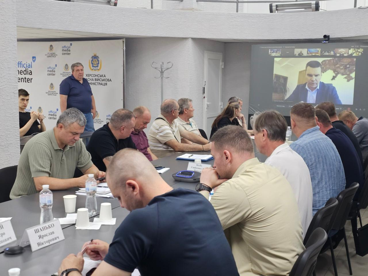 Начальник Херсонської МВА Ярослав Шанько сидить крайнім ліворуч на передньому плані, опустивши голову та роблячи записи в документах, поки інші учасники наради в ХОВА дивляться онлайн-трансляцію виступу на великому екрані.