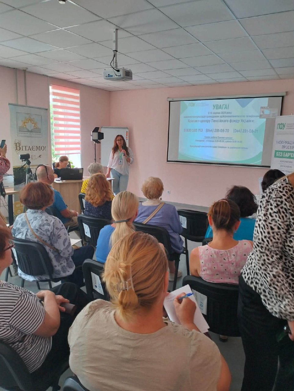 Фахівчиня Пенсійного фонду з мікрофоном у руках виступає перед слухачами у залі бібліотеки ім. Д. Чижевського під час презентації проєкту «Інформаційний простір «БЕЗ БАР'ЄРІВ» для представників Рубанівської громади, демонструючи на великому екрані контакти гарячої лінії ПФУ.