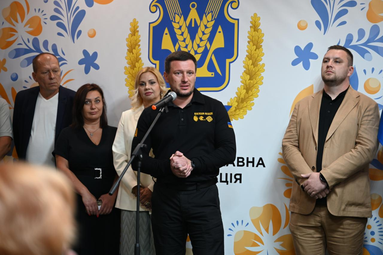 Начальник Херсонської ОВА Олександр Прокудін виступає з мікрофоном на відкритті чотирнадцятого осередку підтримки для жителів Херсонщини в Ужгороді, стоячи поруч із директоркою закарпатського медичного центру «Астра-Діа» Нелею Лазар (ліворуч у білому піджаку), керівником ужгородської стоматологічної клініки LD & Varosh Dental Clinic Олегом Оросом (праворуч у бежевому піджаку) та представниками Херсонської та Закарпатської ОДА.