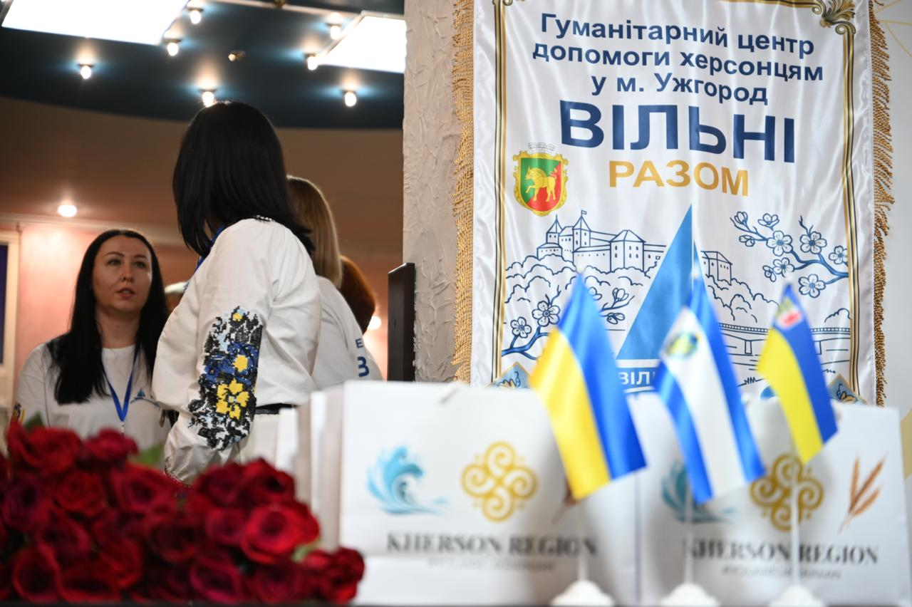 Інтер'єр новоствореного Гуманітарного центру допомоги херсонцям у м. Ужгород «Вільні разом»: на передньому плані — настільні прапорці України та Херсонщини, білі подарункові пакунки «Kherson Region» і букет червоних троянд, праворуч — офіційна хоругва центру, а ліворуч на задньому плані спілкуються працівниці хабу.