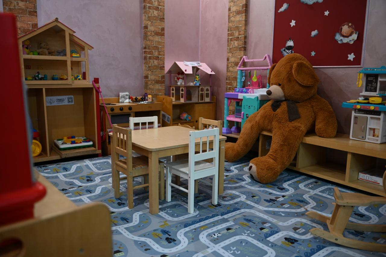 Облаштована дитяча ігрова зона в новому ужгородському гуманітарному центрі «Вільні разом»: по центру стоїть маленький дерев'яний стіл із стільчиками на килимі з автомобільними дорогами, а навколо розміщено полиці з іграшками, лялькові будиночки, дитячу кухню та великого плюшевого ведмедя.