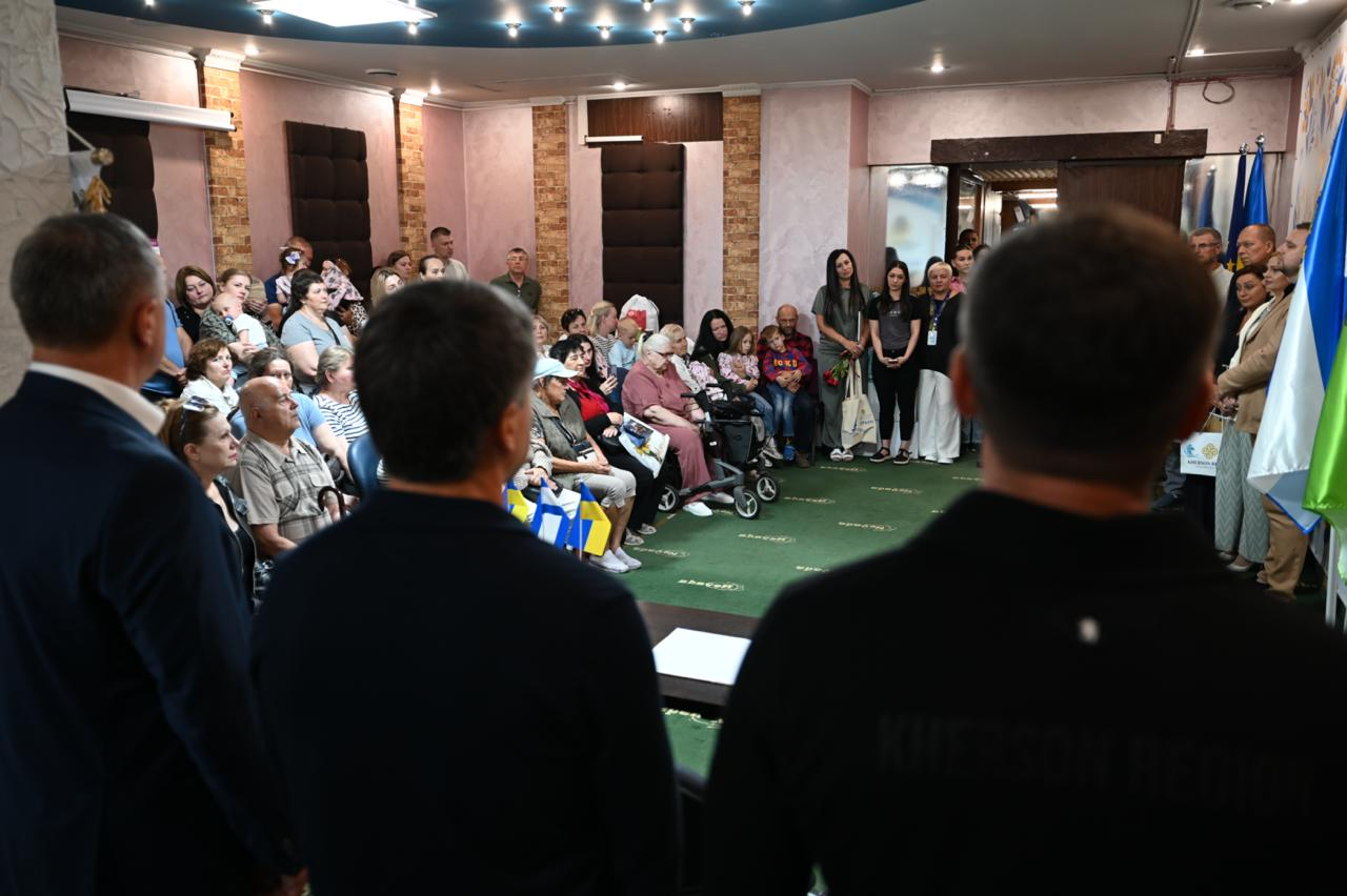 Велика група херсонців-переселенців різного віку сидить та стоїть у залі нового ужгородського центру «Вільні разом» разом із працівниками хабу, слухаючи виступ керівництва Херсонської ОВА та Закарпатської ОДА, що стоять на передньому плані спиною до камери.