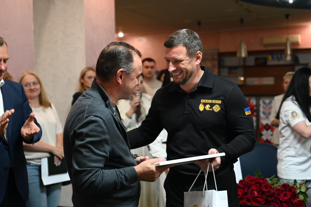 Начальник Херсонської ОВА Олександр Прокудін усміхається та вручає подяку в рамці й пакунок чоловікові в сірому кардигані під час офіційного заходу з нагоди відкриття ужгородського гуманітарного центру «Вільні разом».