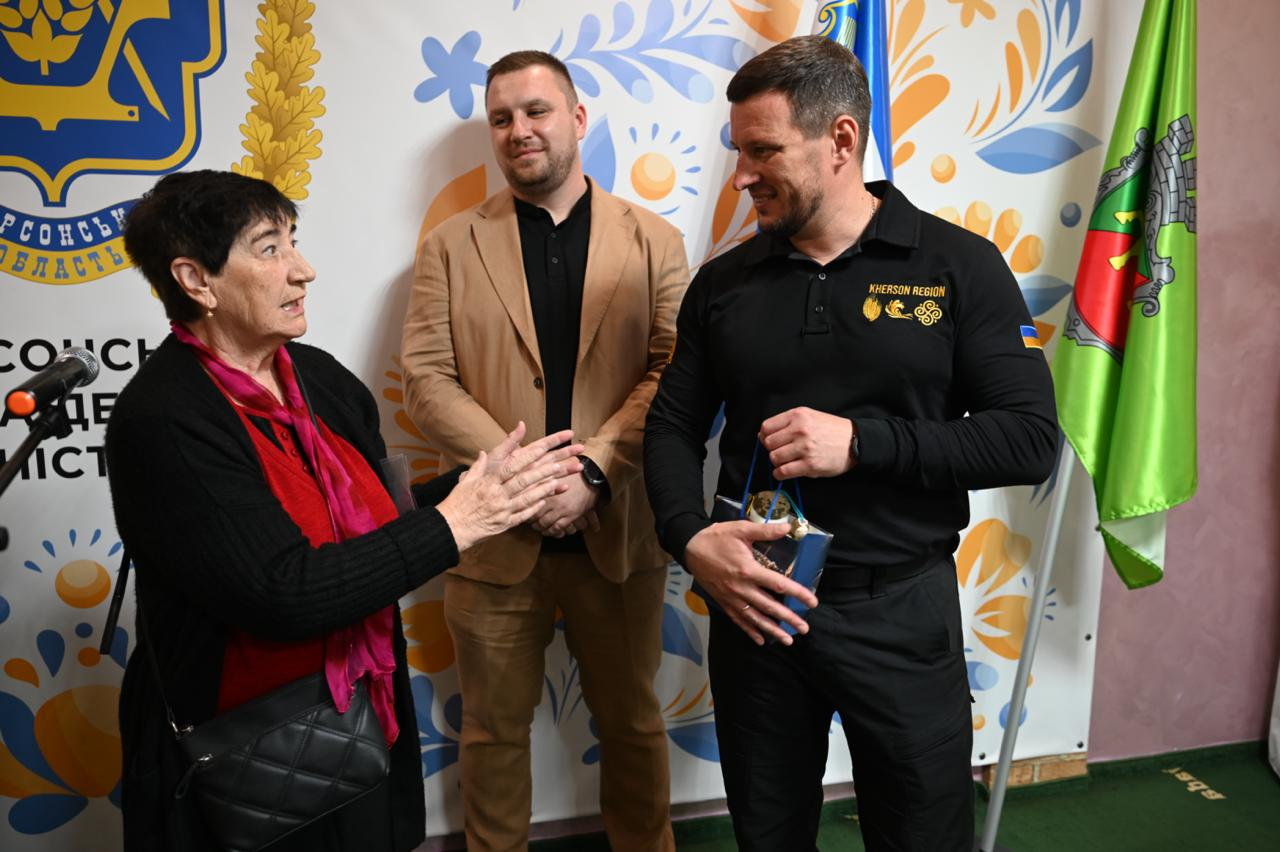 Начальник Херсонської ОВА Олександр Прокудін спілкується з літньою жінкою, яка звернулася до нього, тримаючи в руках подарунок, на тлі офіційного банера хабу та керівника ужгородської клініки LD & Varosh Dental Clinic Олега Ороса (посередині).