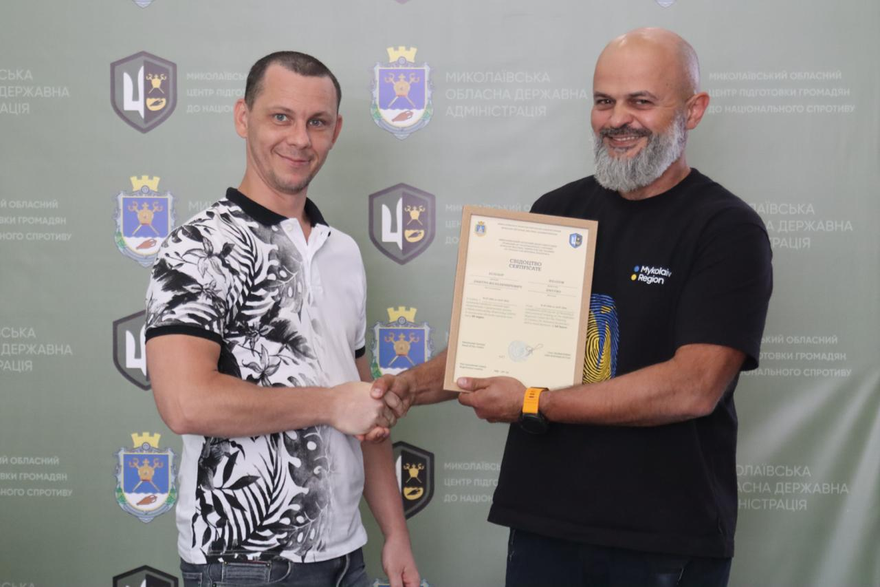 Інструктор Миколаївського обласного центру підготовки громадян до національного спротиву тисне руку та вручає свідоцтво про закінчення курсу базової військової підготовки учаснику вишколу в біло-чорній футболці на тлі офіційного банера.