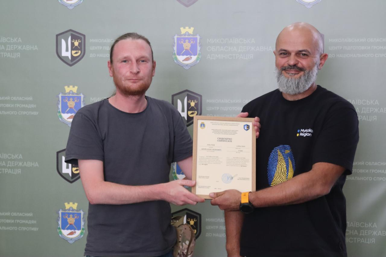 Інструктор Миколаївського обласного центру підготовки громадян до національного спротиву вручає свідоцтво про закінчення курсу базової військової підготовки учаснику вишколу в темно-сірій футболці на тлі офіційного банера.