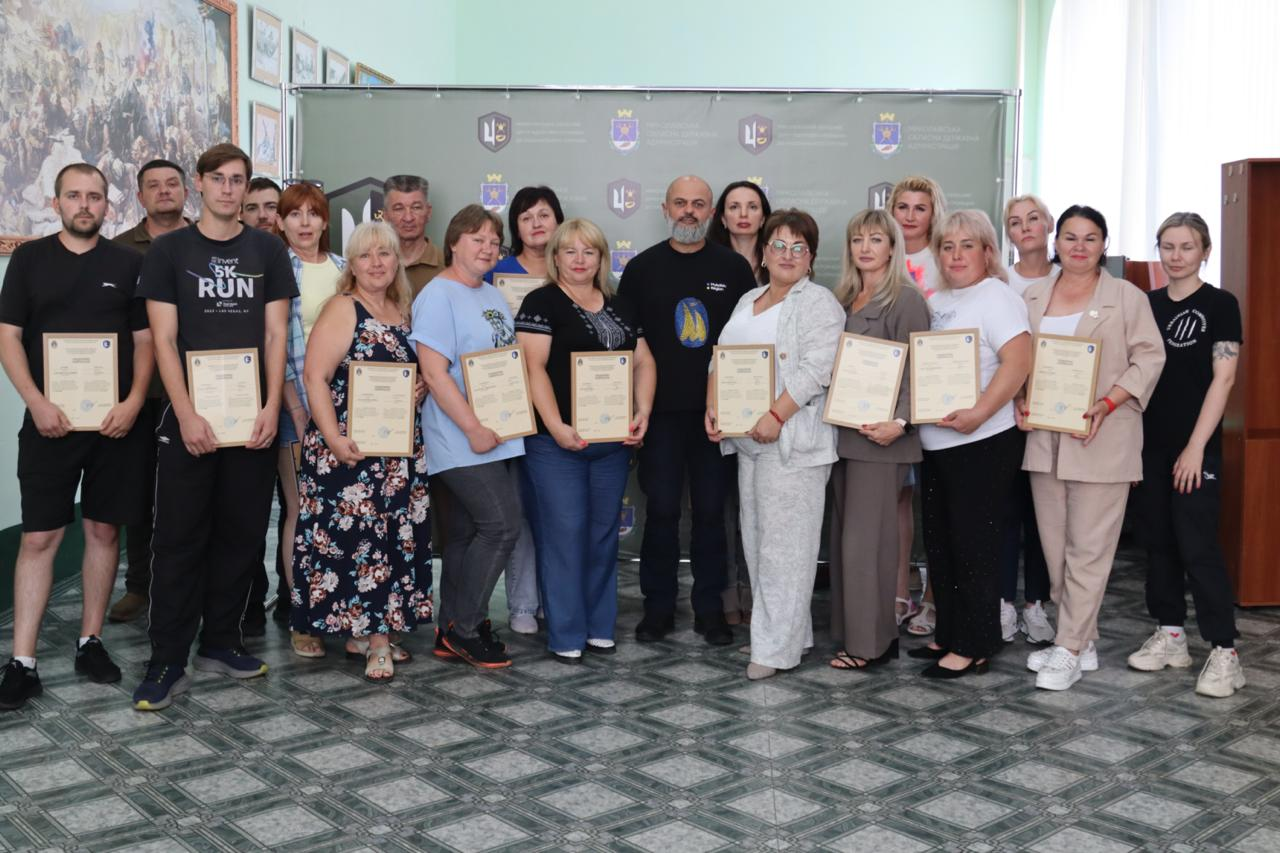 Групове фото іншої частини учасників 23-ї групи фахівців Херсонщини, які тримають у руках свої свідоцтва про закінчення курсу базової військової підготовки разом із головним інструктором по центру на тлі офіційного банера установи.