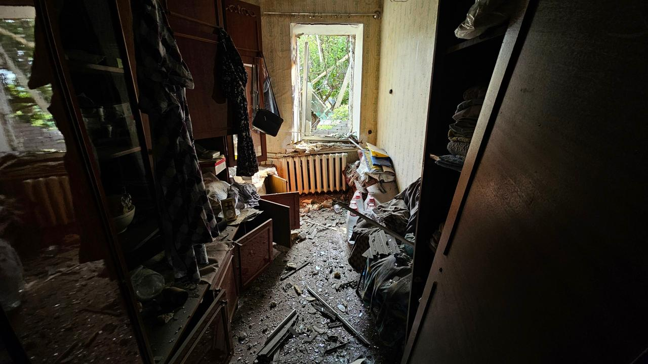Зруйнована внаслідок обстрілу житлова кімната з вибитим вікном, пошкодженими меблями та розкиданими уламками і речами на підлозі.