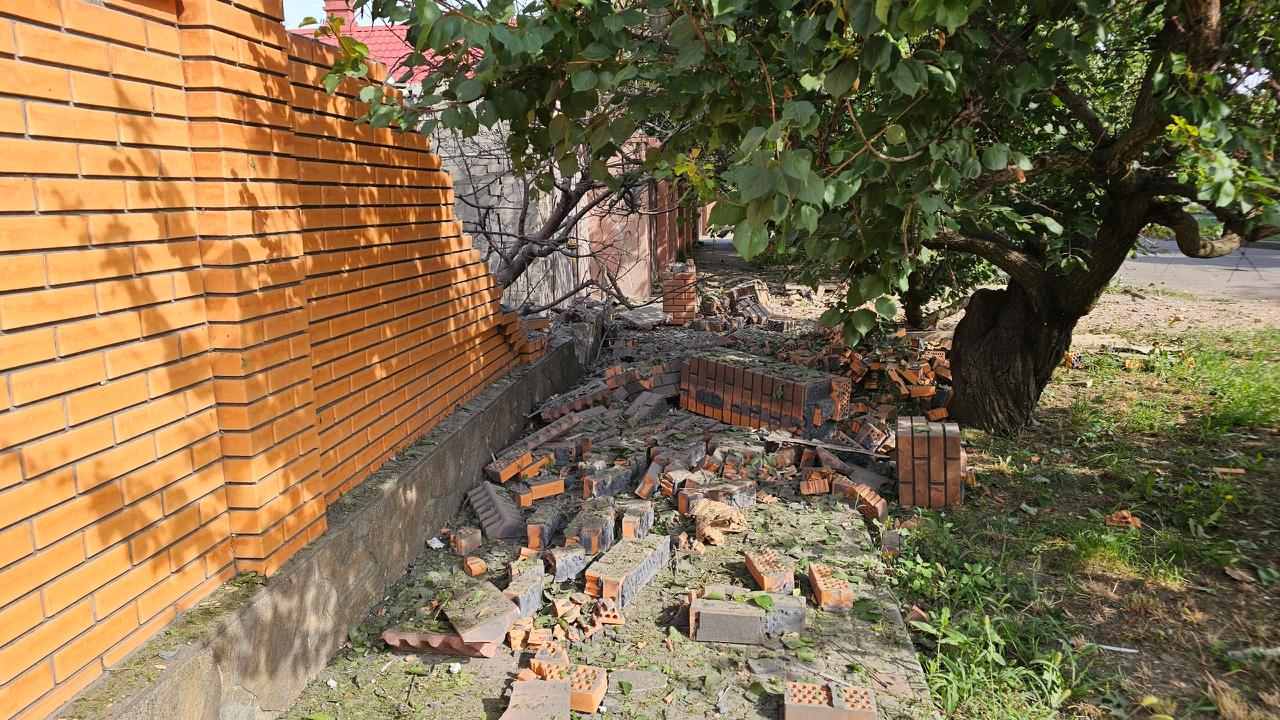 Частково зруйнований цегляний паркан із купою розкиданої цегли та уламків на землі біля дерев.