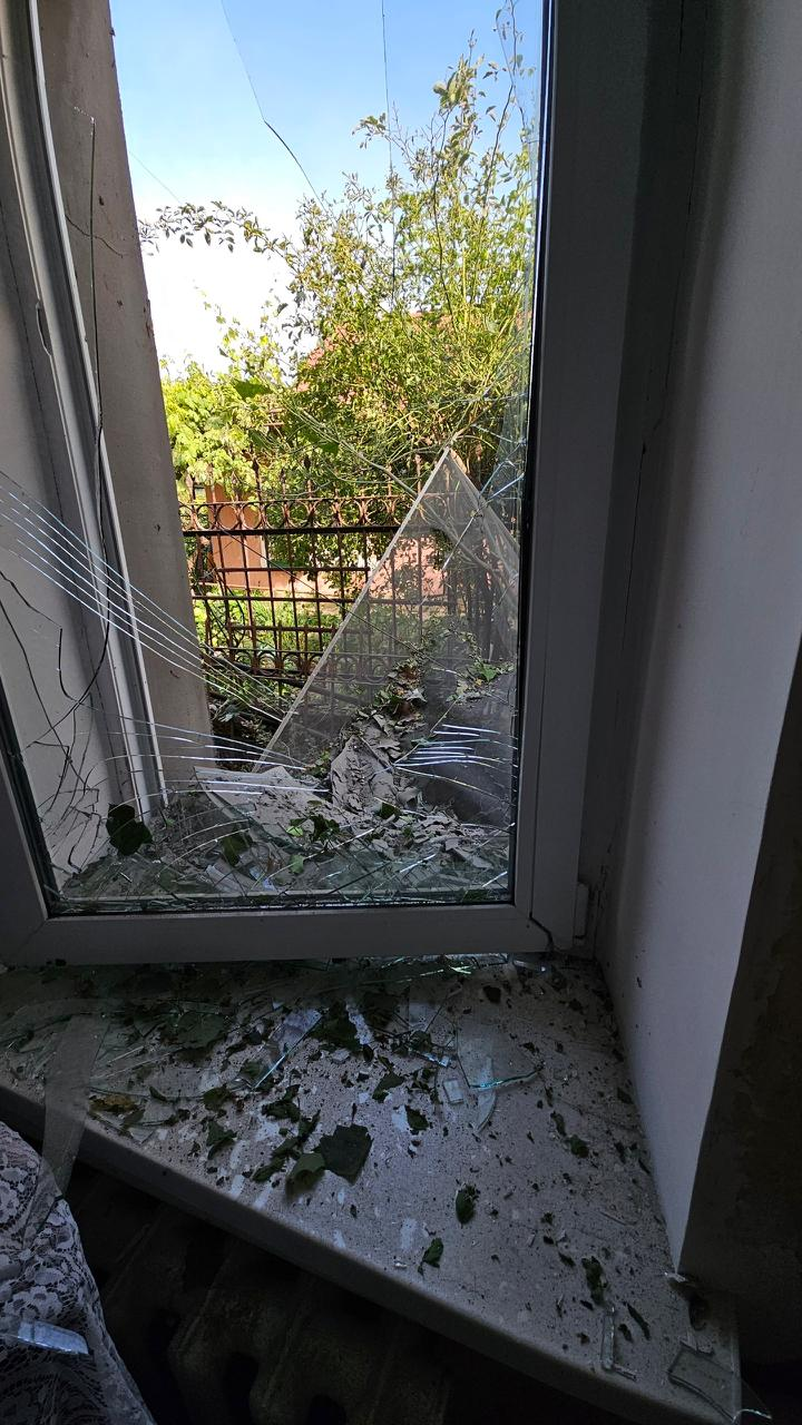 Розбите внаслідок обстрілу металопластикове вікно з потрісканим склом, уламками та залишками листя на підвіконні.