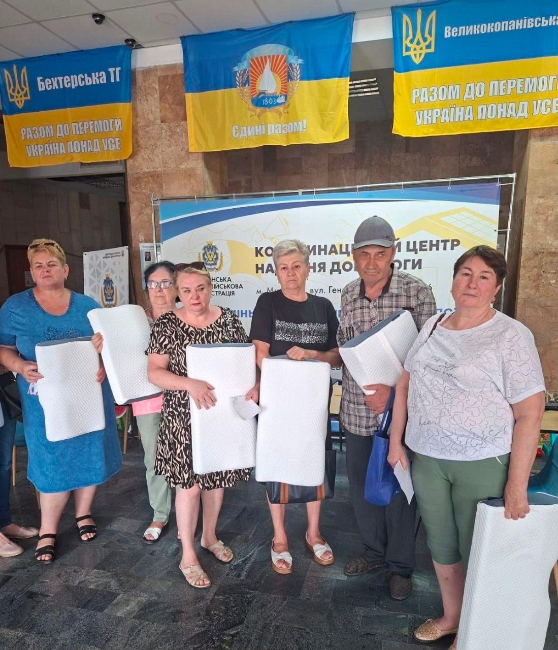 У координаційному центрі «Вільні разом» у Миколаєві внутрішньо переміщені особи з Херсонщини стоять із новими білими ортопедичними подушками, які вони отримали як гуманітарну допомогу. На задньому плані розміщені українські прапори та офіційний банер центру.