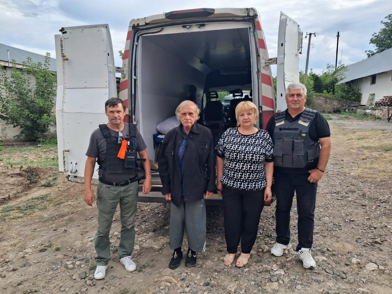Пан Юрій разом із представниками Бериславської громади та волонтерами стоїть на вулиці біля відчинених дверей евакуаційного білого мікроавтобуса. Чоловіки-супроводжуючі одягнені у захисні бронежилети, а вся група готова до виїзду з небезпечної зони.