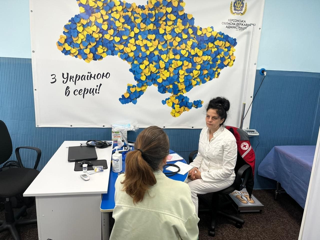 Лікарка мобільної бригади №1 Товариства Червоного Хреста України у білому халаті консультує пацієнтку за робочим столом під час безоплатного прийому ВПО з Херсонщини. На стіні позаду них висить великий банер Херсонської ОДА із картою України, викладеною з синьо-жовтих сердець, та написом «З Україною в серці!».