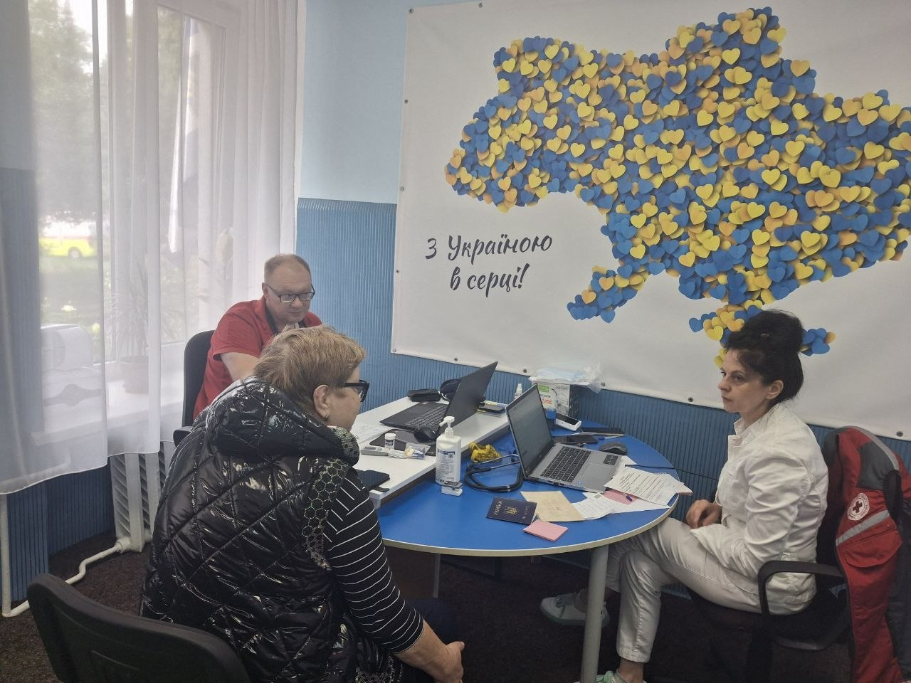 Лікарка мобільної бригади №1 Товариства Червоного Хреста України у білому халаті та чоловік у червоній футболці за круглим столом консультують жінку в чорному жилеті. На столі лежать документи, паспорт та відкриті ноутбуки, а позаду на стіні висить банер із картою України з синьо-жовтих сердець та написом «З Україною в серці!».