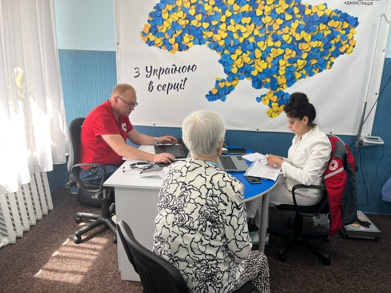Лікарка у білому халаті та лікар у червоній футболці з емблемою Червоного Хреста консультують літню пацієнтку в чорно-білій блузі. Медики працюють за столом із ноутбуками та документами, а на стіні позаду них висить банер із картою України з синьо-жовтих сердець та написом «З Україною в серці!».
