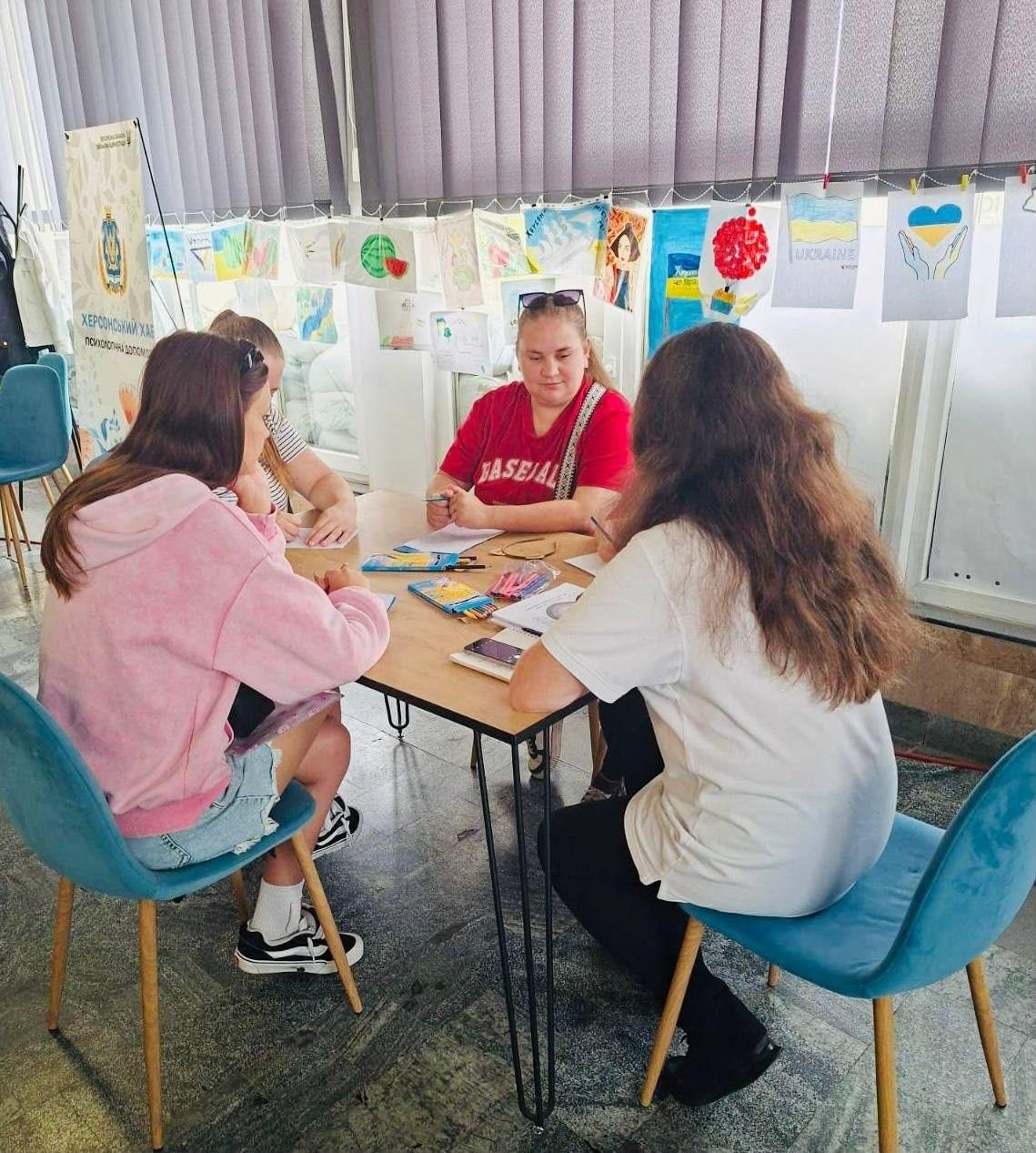 Учасниці творчого майстер-класу від ГО «Дівчата» та координаційного центру «Вільні разом» у Миколаєві сидять за столом, малюють на полотні та спілкуються. На задньому плані видно велике вікно з вертикальними жалюзі, виставку патріотичних малюнків та інформаційний банер «Херсонський хаб».