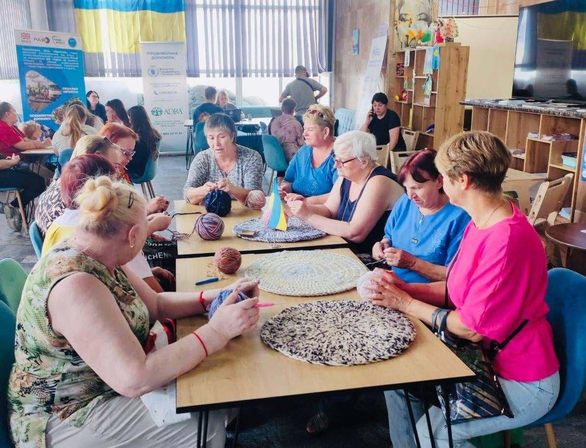 Учасниці гуртка «Коло тепла» координаційного центру «Вільні разом» у Миколаєві сидять за довгим столом у світлому приміщенні хабу, спілкуються та плетуть круглі килимки з кольорової пряжі. На задньому плані видно велике вікно з жалюзі, прапор України та інформаційні банери організацій.