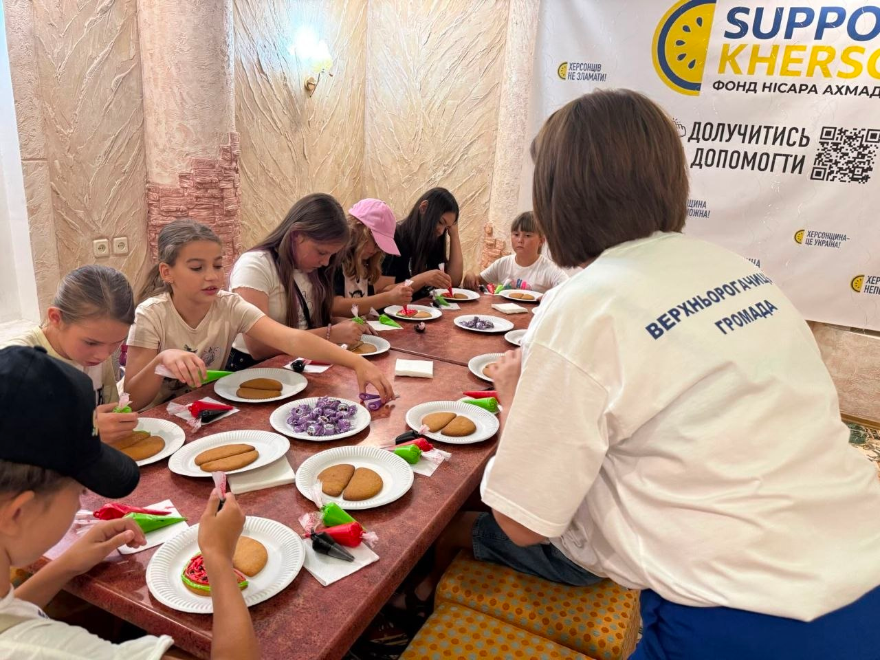 Діти та дорослі в координаційному центрі «Вільні разом» в Одесі сидять за довгим столом на майстер-класі від Верхньорогачицької СВА та розписують кольоровою глазур'ю медові пряники у формі кавунів. На передньому плані видно жінку у білій футболці з написом «Верхньорогачицька громада», а на задньому плані розміщено інформаційний банер фонду «Support Kherson».