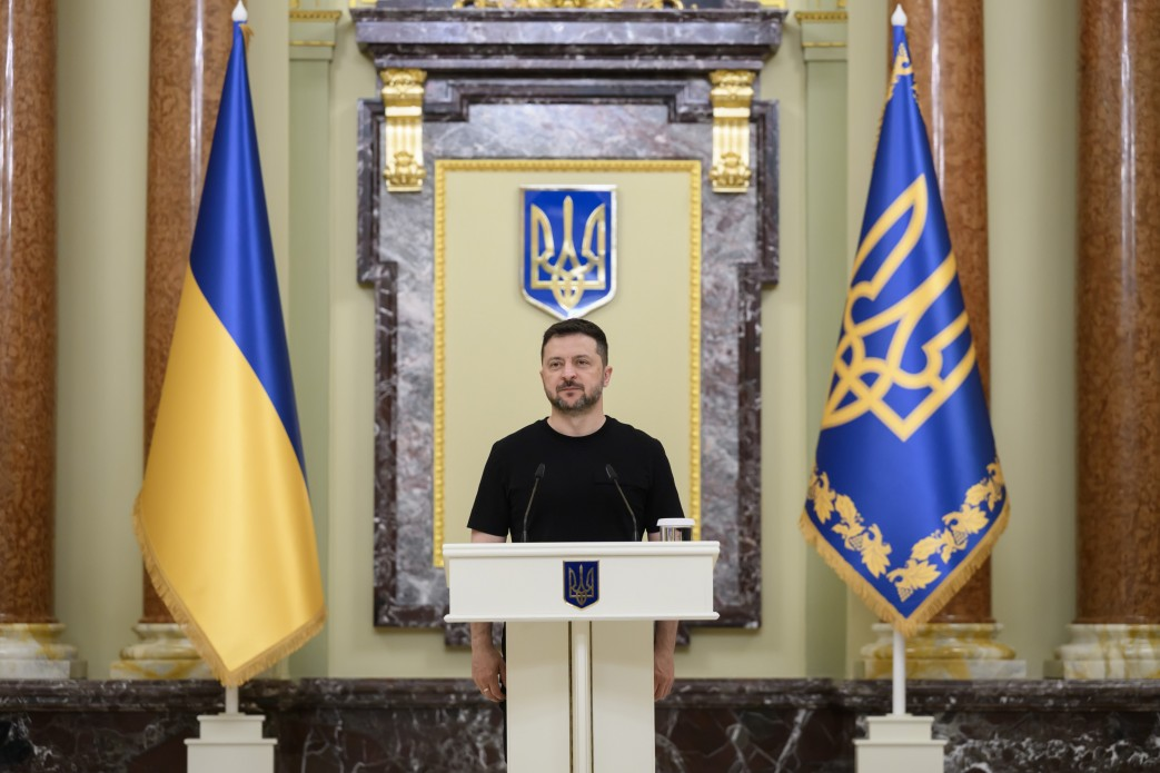 Президент України Володимир Зеленський виступає за білою трибуною на тлі державних прапорів під час зустрічі з бойовими й цивільними медиками напередодні Дня медичних працівників.