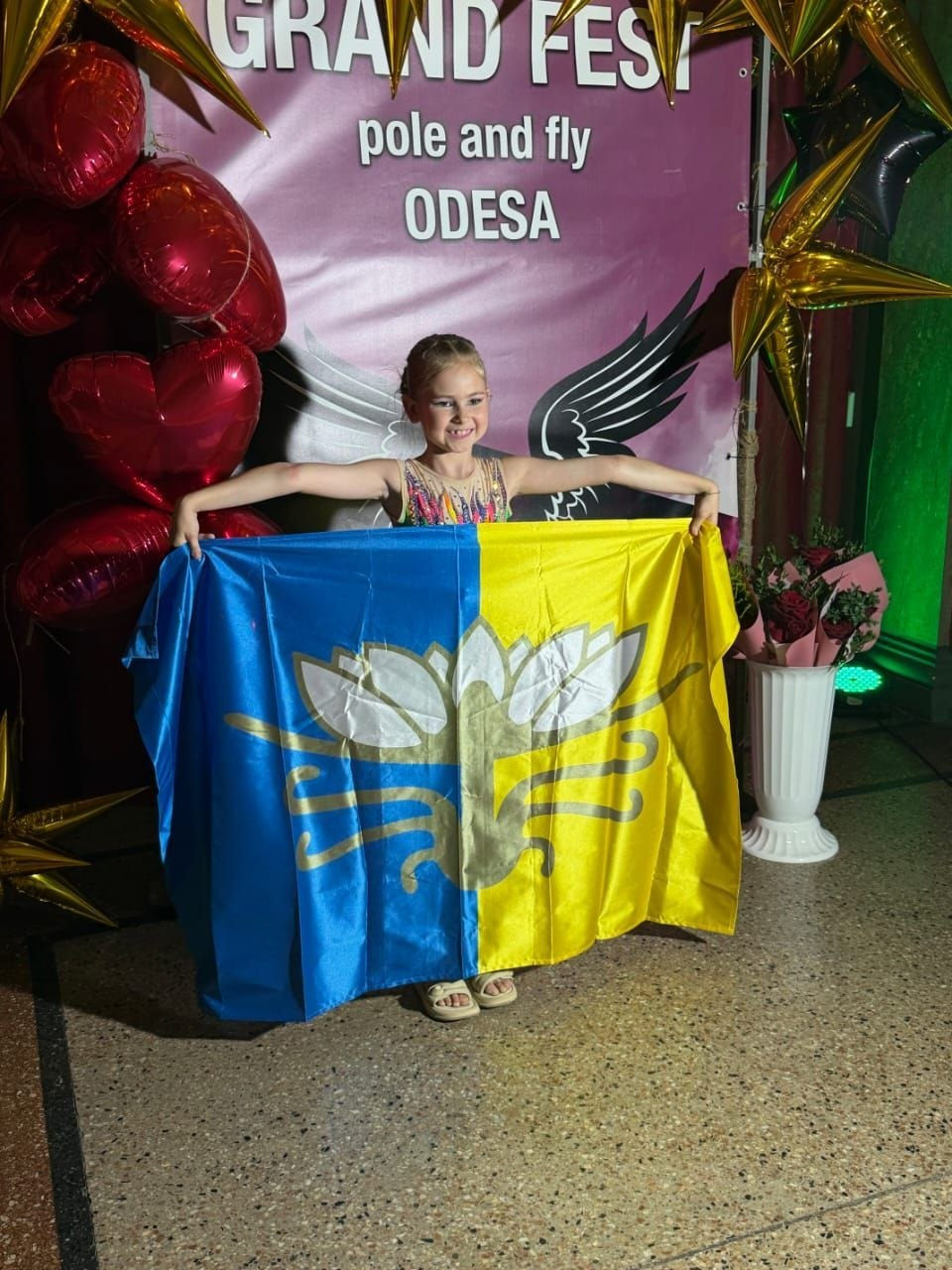 Юна акробатка з Голої Пристані Поліна Болгаріна, переможниця всеукраїнських змагань «Grand Fest» в Одесі, радісно усміхається та тримає синьо-жовтий прапор із золотистим логотипом на тлі святкової фотозони з кульками.