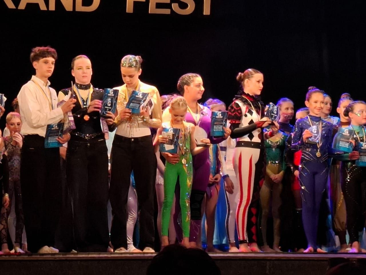 Переможці та призери всеукраїнських змагань «Grand Fest» стоять на сцені під час нагородження, тримаючи в руках кубки, дипломи та медалі, серед яких у центрі — юна акробатка Поліна Болгаріна в яскраво-зеленому костюмі.