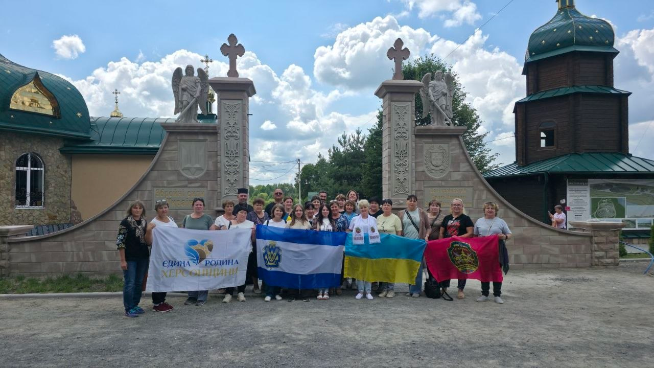 Група учасниць Всеукраїнської прощі — родини загиблих Захисників і Захисниць України — стоять разом біля кам'яної брами духовного комплексу під час поїздки, організованої Херсонською ОДА, та тримають національний прапор України, прапор Херсона, прапор організації «Єдина родина Херсонщини» та червоно-чорний стяг із емблемою.