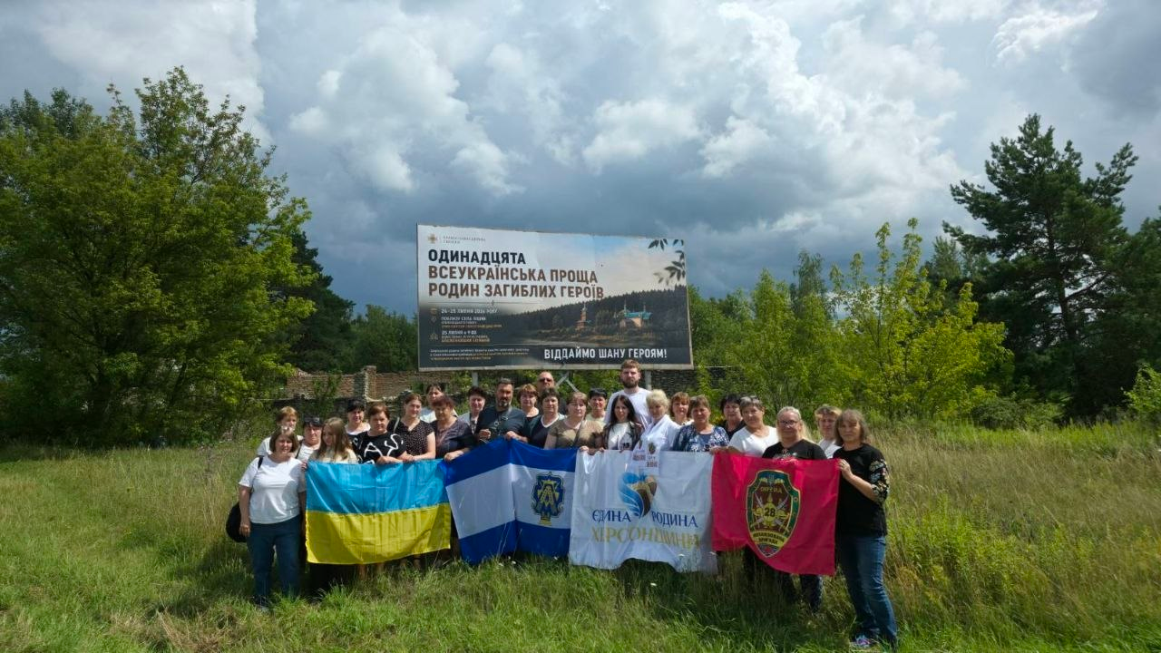 Група учасників Одинадцятої Всеукраїнської прощі родин загиблих Героїв стоїть на галявині біля великого інформаційного банера заходу та тримає державний прапор України, прапор Херсона, стяг організації «Єдина родина Херсонщини» і малиновий прапор із військовою символікою.