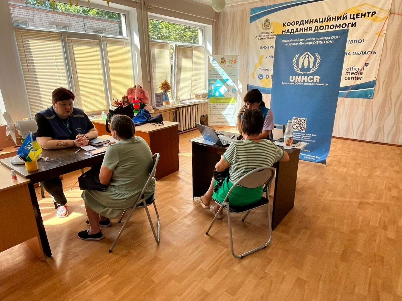 Працівники координаційного центру «Вільні разом» у Кривому Розі ведуть прийом внутрішньо переміщених осіб із Херсонщини за двома робочими столами на тлі великого банера Агентства ООН у справах біженців (UNHCR).