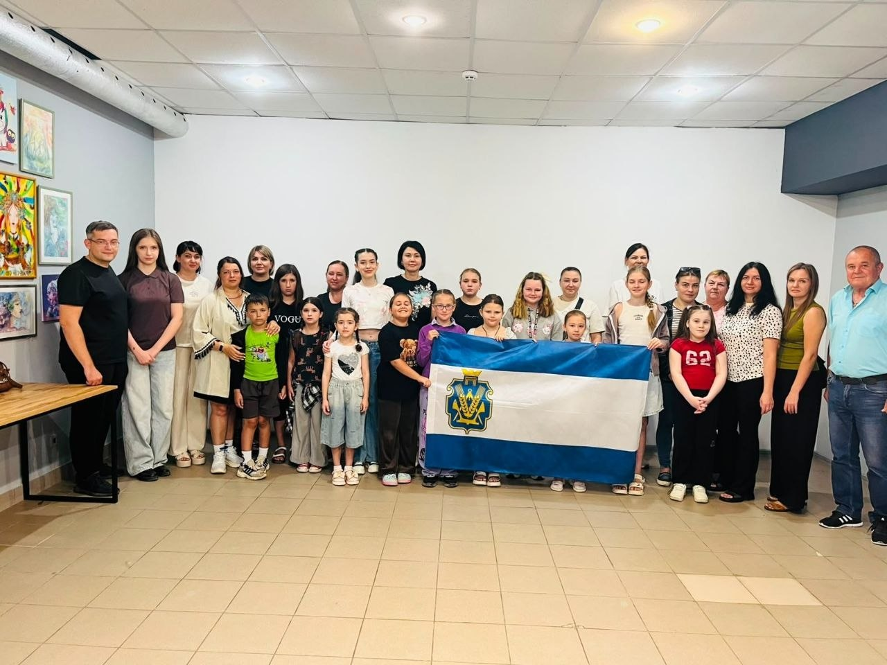Велика група дорослих та дітей — переселенців із Херсонщини в Кривому Розі — стоїть разом у залі під час зустрічі в координаційному центрі «Вільні разом», а діти в центрі тримають синьо-біло-синій прапор Херсонської області з гербом.