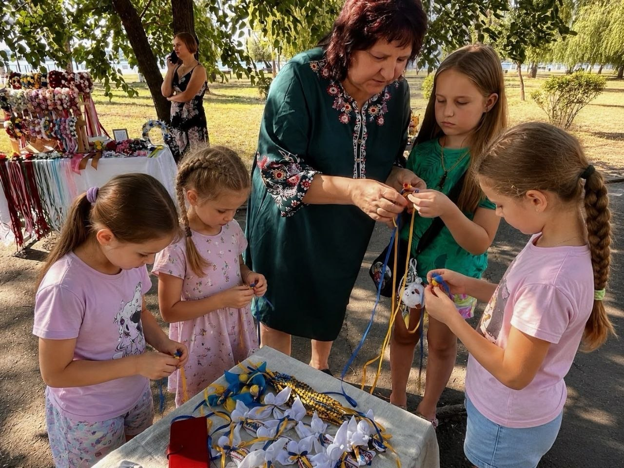 Доросла жінка у вишиванці допомагає дітям-переселенцям із Херсонщини на творчому майстер-класі під час благодійного ярмарку в Миколаєві, де вони разом виготовляють патріотичні сувеніри із синьо-жовтих стрічок для збору коштів на підтримку Сил оборони.