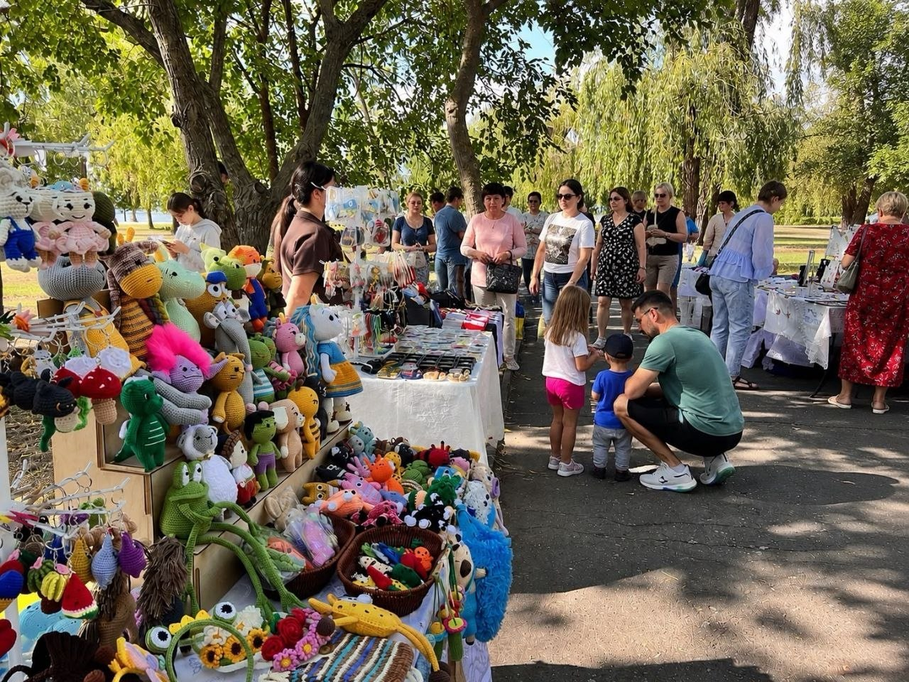 Відвідувачі благодійного ярмарку в Миколаєві розглядають вироби ручної роботи, серед яких на передньому плані — великий стенд із різнокольоровими в'язаними іграшками та сувенірами, створеними для збору коштів на підтримку Сил оборони.